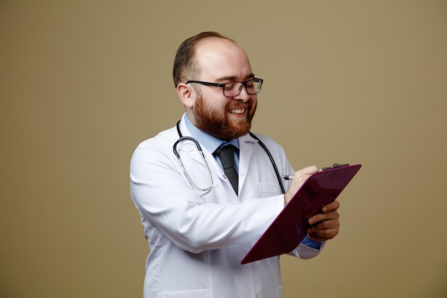 Улыбающийся молодой врач-мужчина в очках, лабораторном халате и стетоскопе на шее пишет в буфер обмена ручкой, изолированной на оливково-зеленом фоне