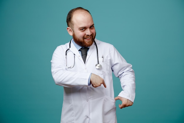 Foto gratuita sorridente giovane medico maschio che indossa cappotto medico e stetoscopio intorno al collo guardando la fotocamera che punta alla sua tasca isolata su sfondo blu