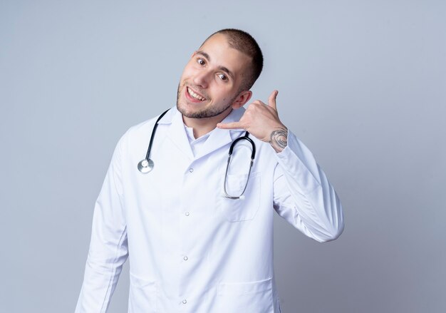 흰 벽에 고립 된 전화 제스처를 하 고 그의 목에 의료 가운과 청진기를 입고 웃는 젊은 남성 의사