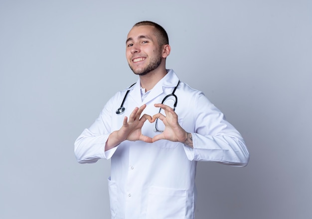 Smiling young male doctor wearing medical robe and stethoscope around his neck doing heart sign isolated on white wall