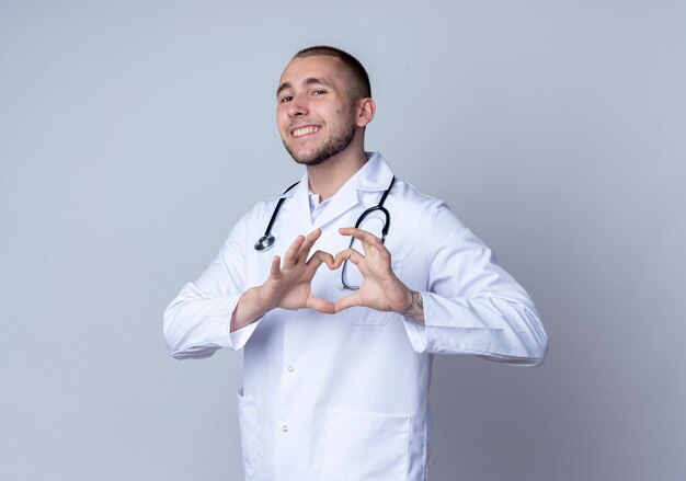 Smiling young male doctor wearing medical robe and stethoscope around his neck doing heart sign isolated on white wall