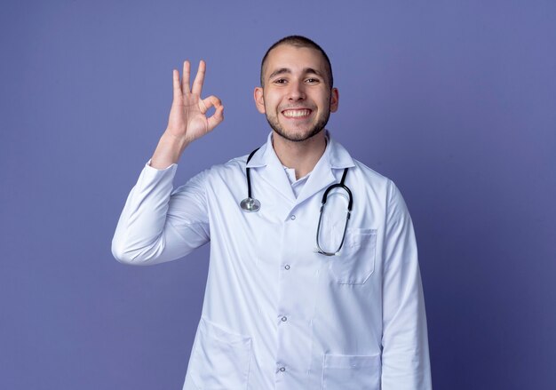 Smiling young male doctor wearing medical robe and stethoscope around his neck doing ok sign isolated on purple wall