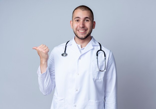 Smiling young male doctor wearing medical robe and stethoscope around his neck pointing at side isolated on white wall