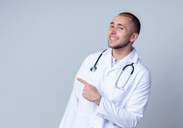 Smiling young male doctor wearing medical robe and stethoscope around his neck pointing at side isolated on white wall