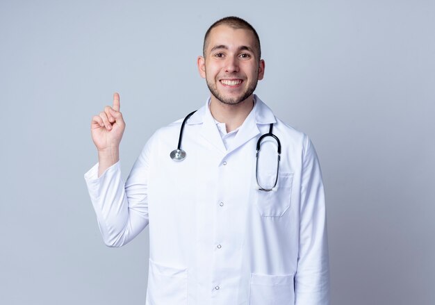 흰 벽에 고립 된 손가락을 올리는 그의 목 주위에 의료 가운과 청진기를 입고 웃는 젊은 남성 의사