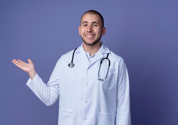 Smiling young male doctor wearing medical robe and stethoscope around his neck showing empty hand isolated on purple wall