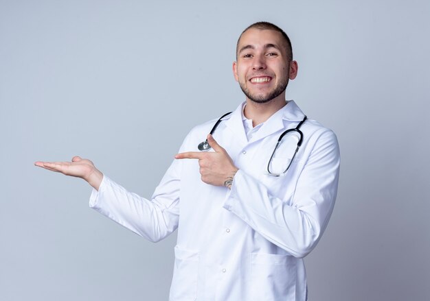 Smiling young male doctor wearing medical robe and stethoscope around his neck showing empty hand and pointing at it isolated on white wall