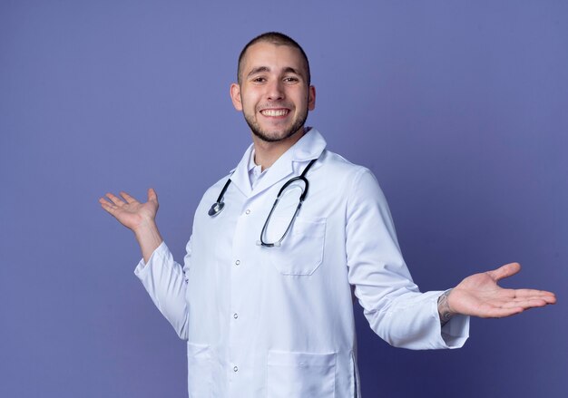 紫色の壁に隔離された空の手を示す彼の首の周りに医療ローブと聴診器を身に着けている若い男性医師の笑顔