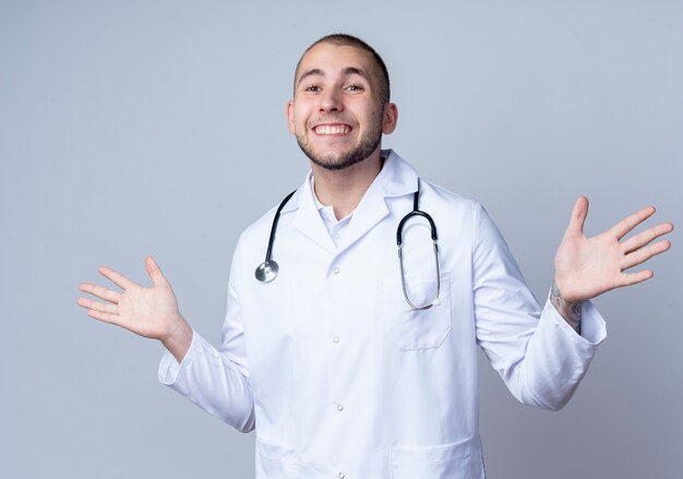 Улыбающийся молодой мужчина-врач в медицинском халате и стетоскопе на шее показывает пустые руки, изолированные на белой стене