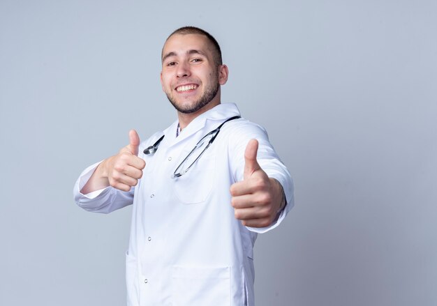 흰 벽에 고립 엄지 손가락을 보여주는 그의 목 주위에 의료 가운과 청진기를 입고 웃는 젊은 남성 의사