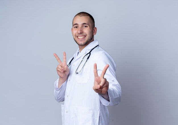 Smiling young male doctor wearing medical robe and stethoscope around his neck standing in profile view doing peace signs isolated on white wall