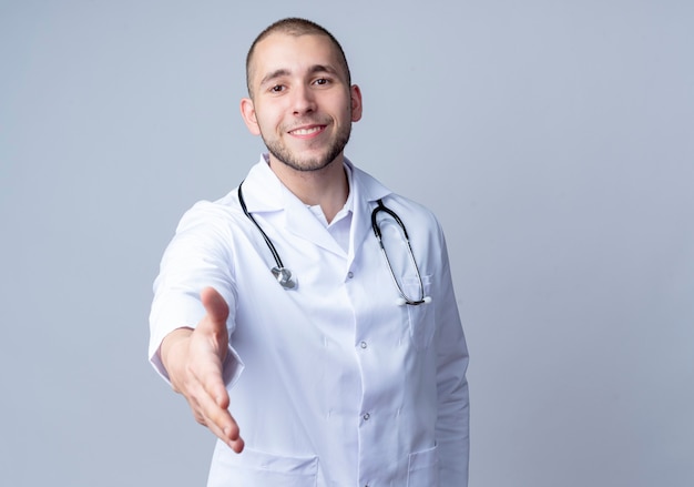 흰 벽에 고립 된 앞에서 몸짓으로 손을 뻗어 그의 목 주위에 의료 가운과 청진기를 입고 웃는 젊은 남성 의사