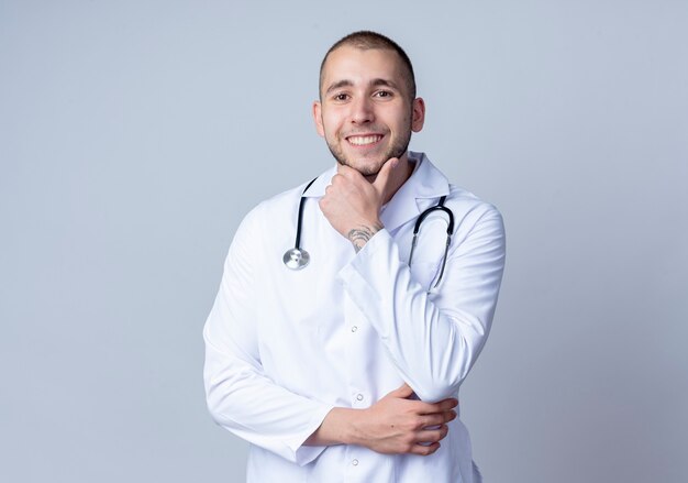 Улыбающийся молодой мужчина-врач в медицинском халате и стетоскопе на шее, касающийся его подбородка, изолированного на белой стене