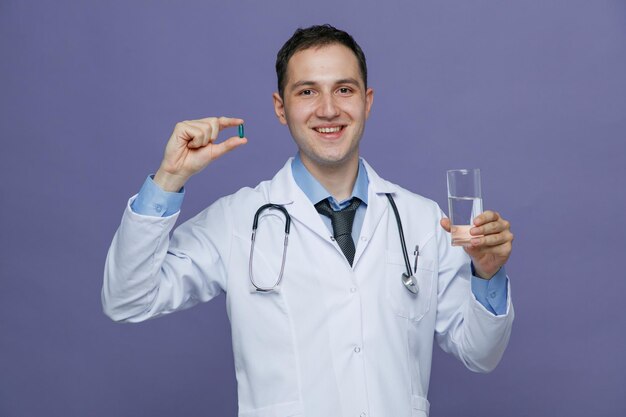 Smiling young male doctor wearing medical robe and stethoscope around neck holding glass of water looking at camera showing capsule isolated on purple background