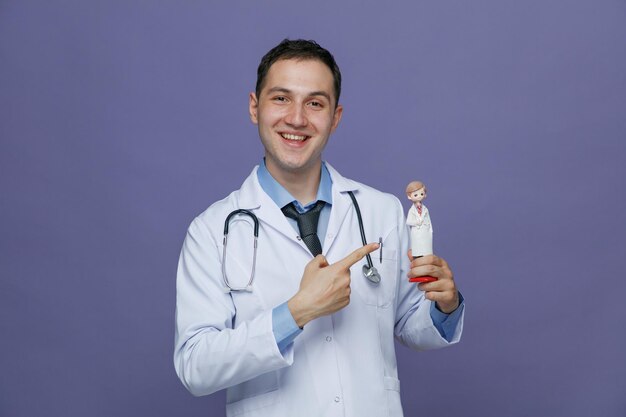 Smiling young male doctor wearing medical robe and stethoscope around neck showing doctor figurine pointing at it looking at camera isolated on purple background