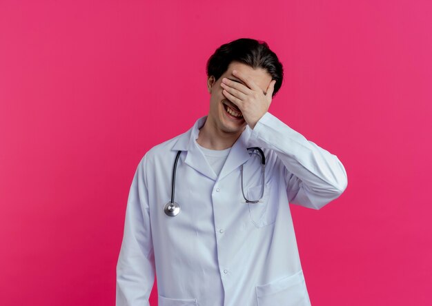 Улыбающийся молодой мужчина-врач в медицинском халате и стетоскопе, закрывающий глаза рукой, изолированной на розовой стене с копией пространства