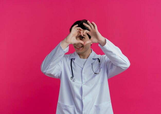 의료 가운과 청진기를 입고 웃는 젊은 남성 의사 복사 공간이 분홍색 벽에 고립 된 심장 기호를 하 고