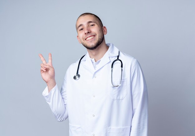 Smiling young male doctor wearing medical robe and stethoscope doing peace sign around his neck isolated on white wall