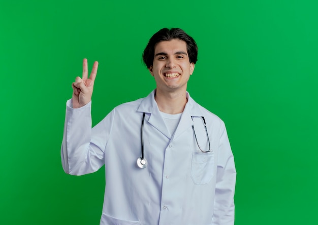 コピースペースと緑の壁に分離されたピースサインをしている医療ローブと聴診器を身に着けている若い男性医師の笑顔