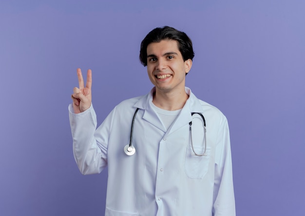 コピースペースと紫色の壁に分離されたピースサインをしている医療ローブと聴診器を身に着けている若い男性医師の笑顔