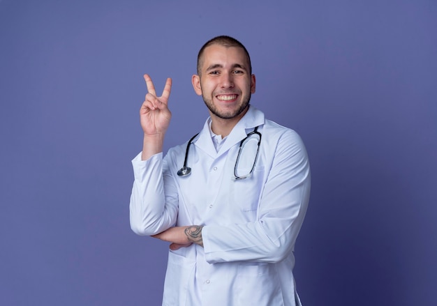 Sorridente giovane medico maschio indossa abito medico e stetoscopio facendo segno di pace mettendo la mano sotto il gomito isolato sulla parete viola