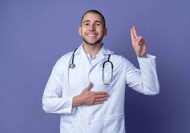 Smiling young male doctor wearing medical robe and stethoscope doing promise gesture isolated on purple wall