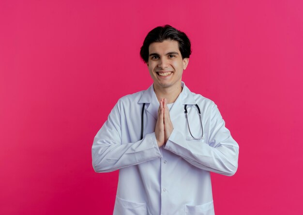 Smiling young male doctor wearing medical robe and stethoscope  keeping hands together isolated on pink wall with copy space