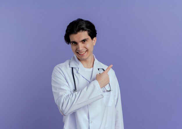 Smiling young male doctor wearing medical robe and stethoscope looking pointing behind isolated