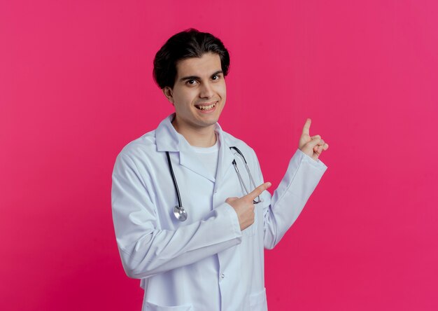 コピースペースとピンクの壁に分離された後ろを指す医療ローブと聴診器を身に着けている若い男性医師の笑顔