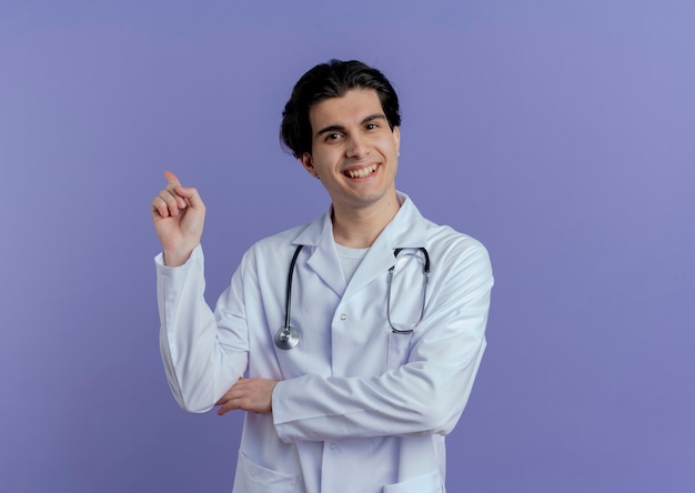 Smiling young male doctor wearing medical robe and stethoscope  pointing behind isolated on purple wall with copy space