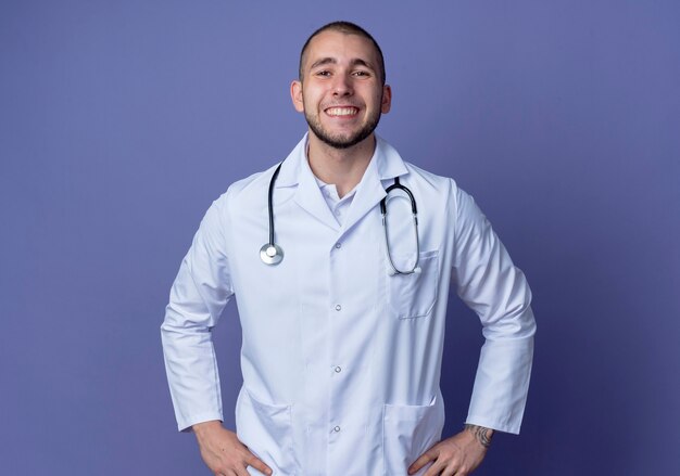 보라색 벽에 고립 된 허리에 손을 댔을 의료 가운과 청진기를 입고 웃는 젊은 남성 의사