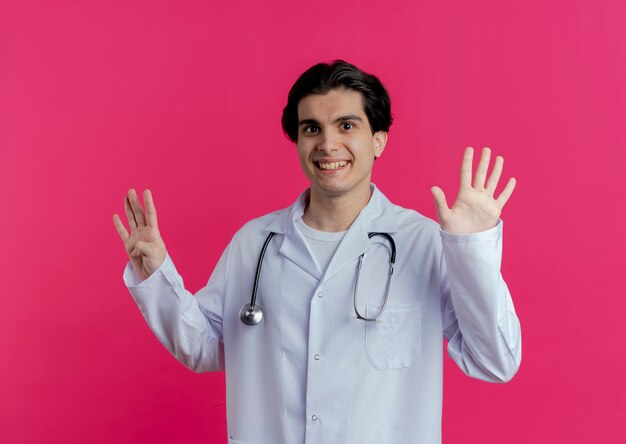 ピンクの壁に隔離された手で9を示す医療ローブと聴診器を身に着けている若い男性医師の笑顔