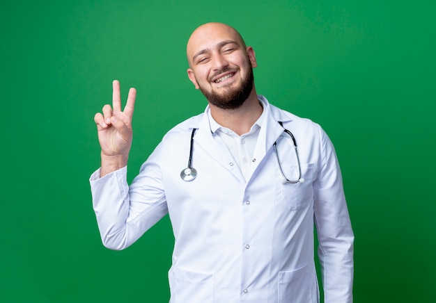 Smiling young male doctor wearing medical robe and stethoscope showing okey gesture isolated on green