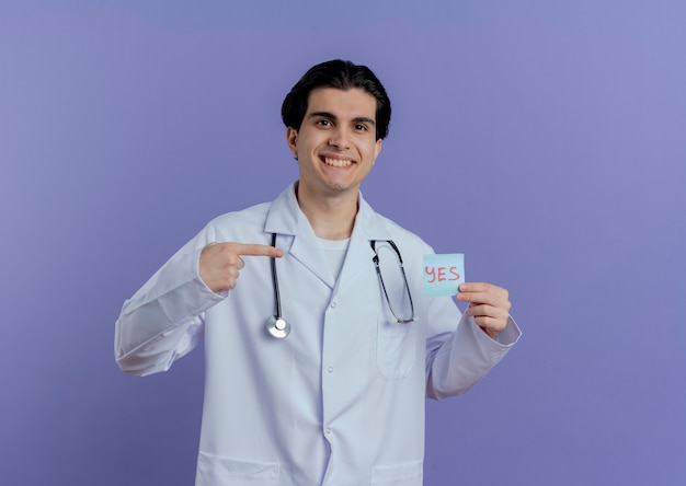 의료 가운과 청진기를 입고 웃는 젊은 남성 의사 그것을 가리키는 예 참고 및 복사 공간이 보라색 벽에 고립