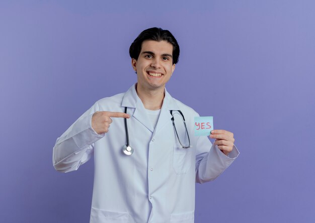 Smiling young male doctor wearing medical robe and stethoscope showing yes note pointing at it and  isolated on purple wall with copy space