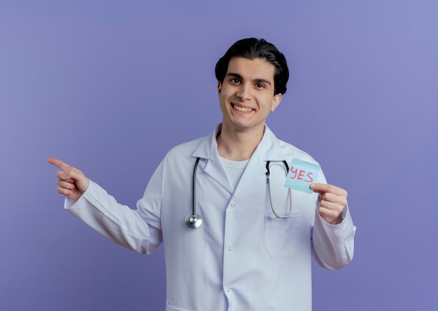 Улыбающийся молодой мужчина-врач в медицинском халате и стетоскопе показывает примечание да, указывающее на сторону, изолированную на фиолетовой стене с копией пространства