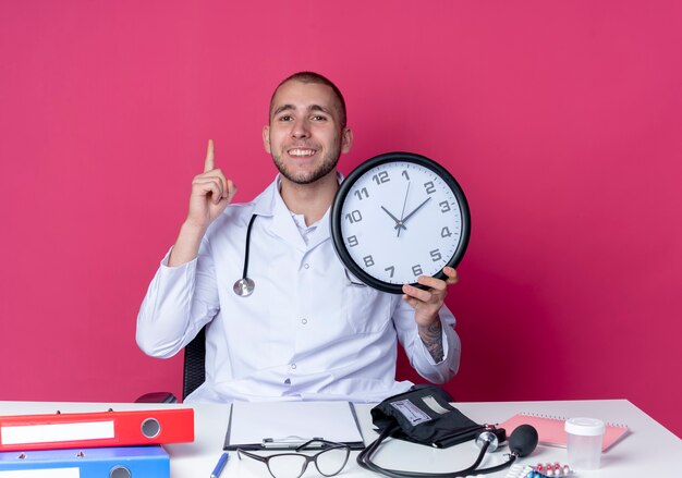 Smiling young male doctor wearing medical robe and stethoscope sitting at desk with work tools holding clock and raising finger isolated on pink wall