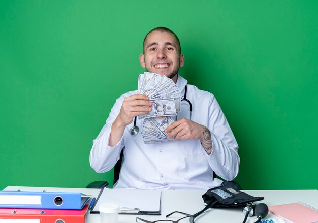 緑の壁に隔離されたお金を保持している作業ツールと机に座って医療ローブと聴診器を身に着けている若い男性医師の笑顔