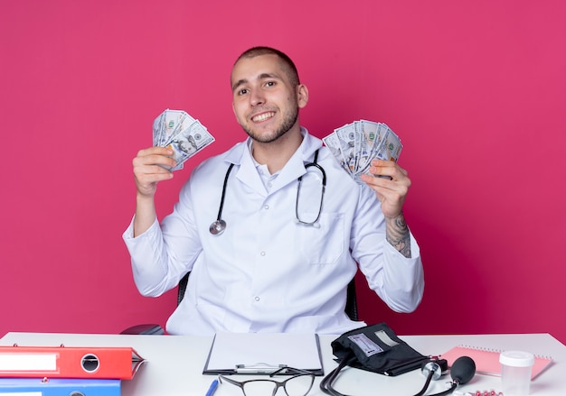 Foto gratuita sorridente giovane medico maschio indossa abito medico e stetoscopio seduto alla scrivania con strumenti di lavoro che tengono soldi isolati sulla parete rosa