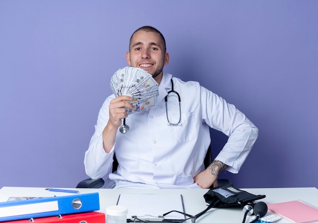 보라색 벽에 고립 된 허리에 손으로 돈을 들고 작업 도구로 책상에 앉아 의료 가운과 청진기를 입고 웃는 젊은 남성 의사