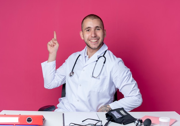 Smiling young male doctor wearing medical robe and stethoscope sitting at desk with work tools pointing up isolated on pink wall