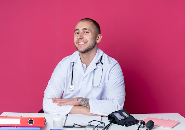 Улыбающийся молодой мужчина-врач в медицинском халате и стетоскопе сидит за столом с рабочими инструментами, положив руки на стол, изолированный на розовой стене