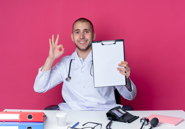 Smiling young male doctor wearing medical robe and stethoscope sitting at desk with work tools showing clipboard and doing ok sign isolated on pink wall