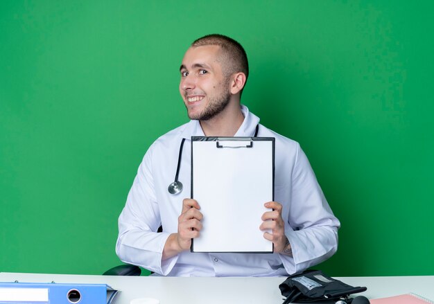 Smiling young male doctor wearing medical robe and stethoscope sitting at desk with work tools showing clipboard isolated on green wall