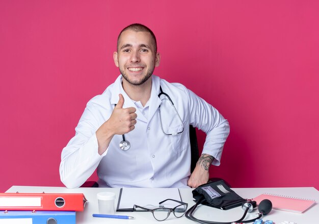 의료 가운과 청진기를 입고 웃는 젊은 남성 의사가 분홍색 벽에 고립 된 엄지 손가락을 보여주는 작업 도구로 책상에 앉아