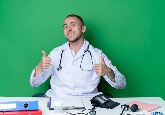 Smiling young male doctor wearing medical robe and stethoscope sitting at desk with work tools showing thumbs up isolated on green wall