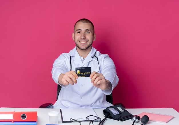 Free photo smiling young male doctor wearing medical robe and stethoscope sitting at desk with work tools stretching out credit card towards front isolated on pink wall