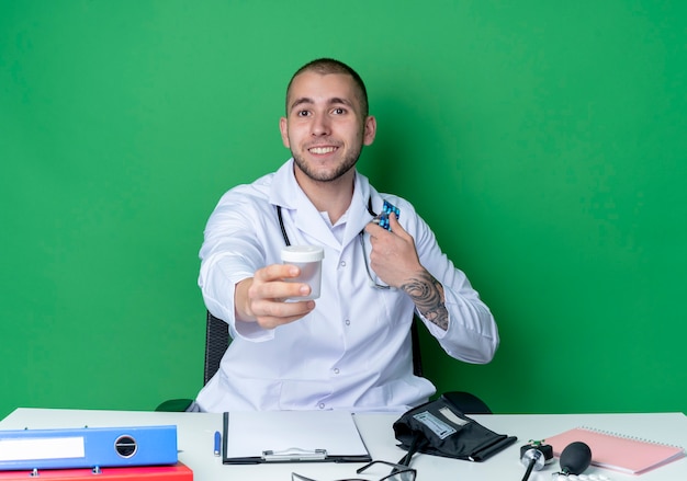 의료 가운과 청진기를 착용하는 젊은 남성 의사가 앞쪽으로 의료 비커를 펴고 녹색 벽에 고립 된 캡슐 팩을 들고 작업 도구로 책상에 앉아 웃고