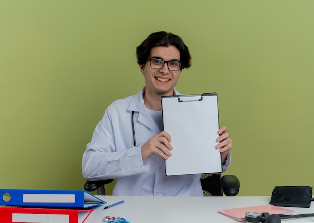 Sorridente giovane medico maschio che indossa abito medico e stetoscopio con gli occhiali seduto alla scrivania con strumenti medici guardando mostrando appunti isolati