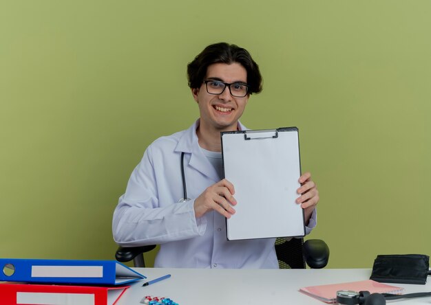 젊은 남성 의사 의료 가운과 청진기를 착용하고 의료 도구를 책상에 앉아 안경을 보여주는 클립 보드 격리를 찾고 웃고